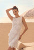 Woman wearing a white lace dress on a beach with sand and an umbrella in the background.