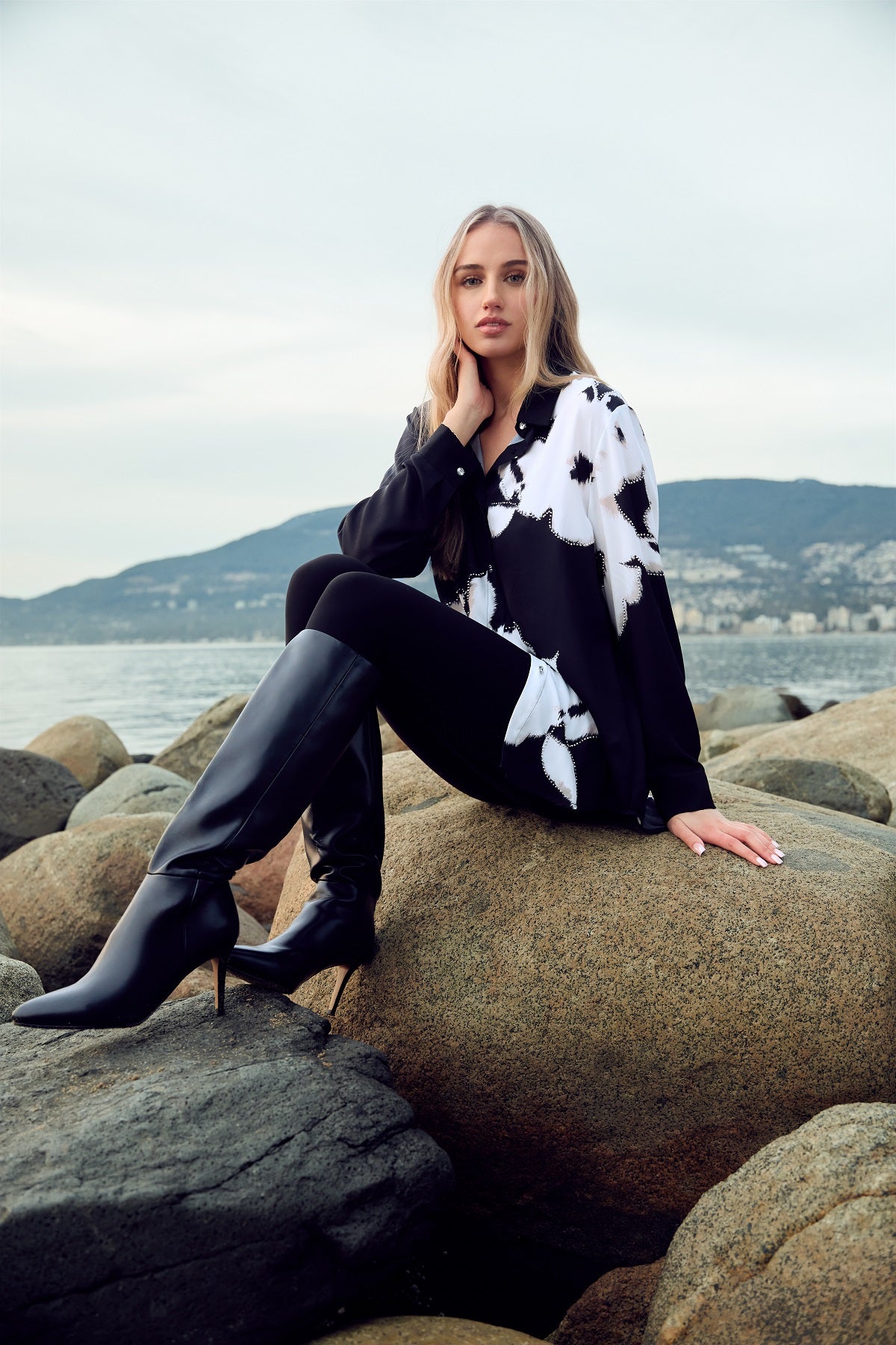 Woman sitting on rocks by the water wearing a black and white patterned Blouse