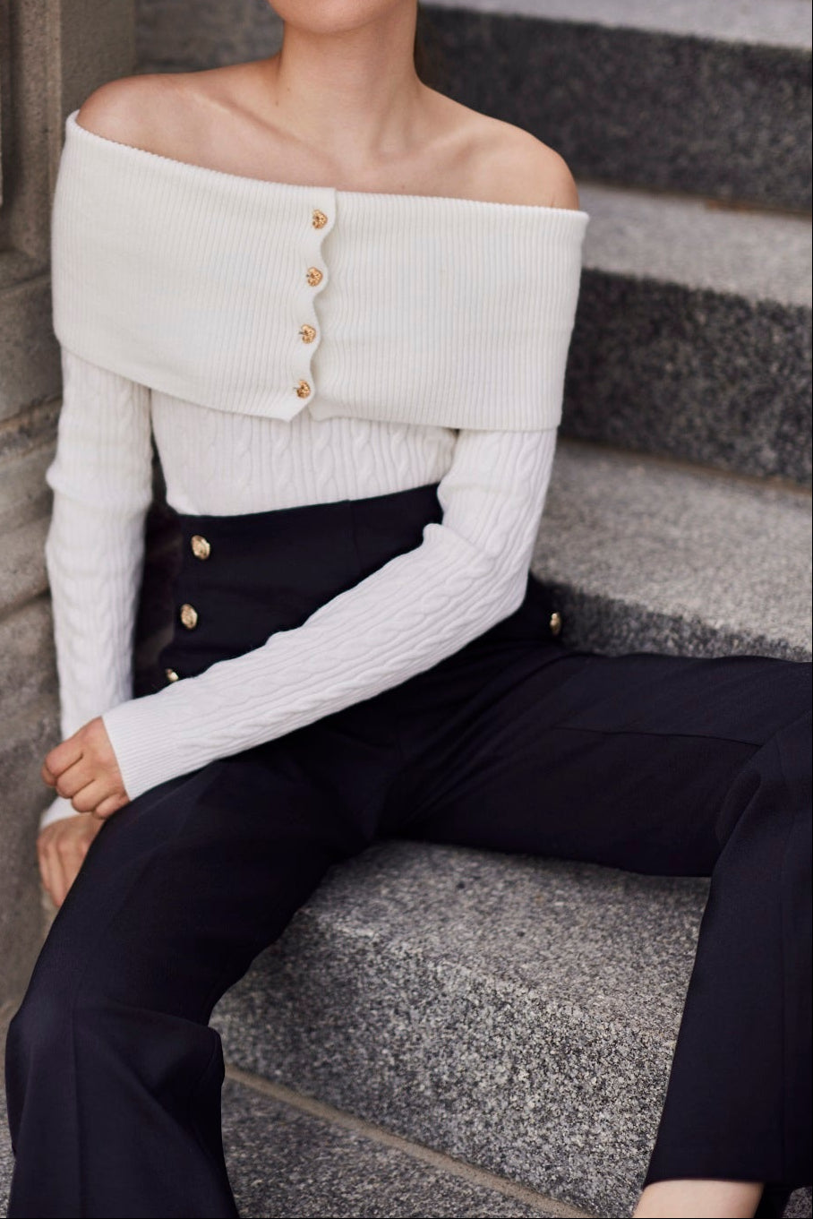 Woman wearing a white off-shoulder top with buttons, sitting on stone steps.