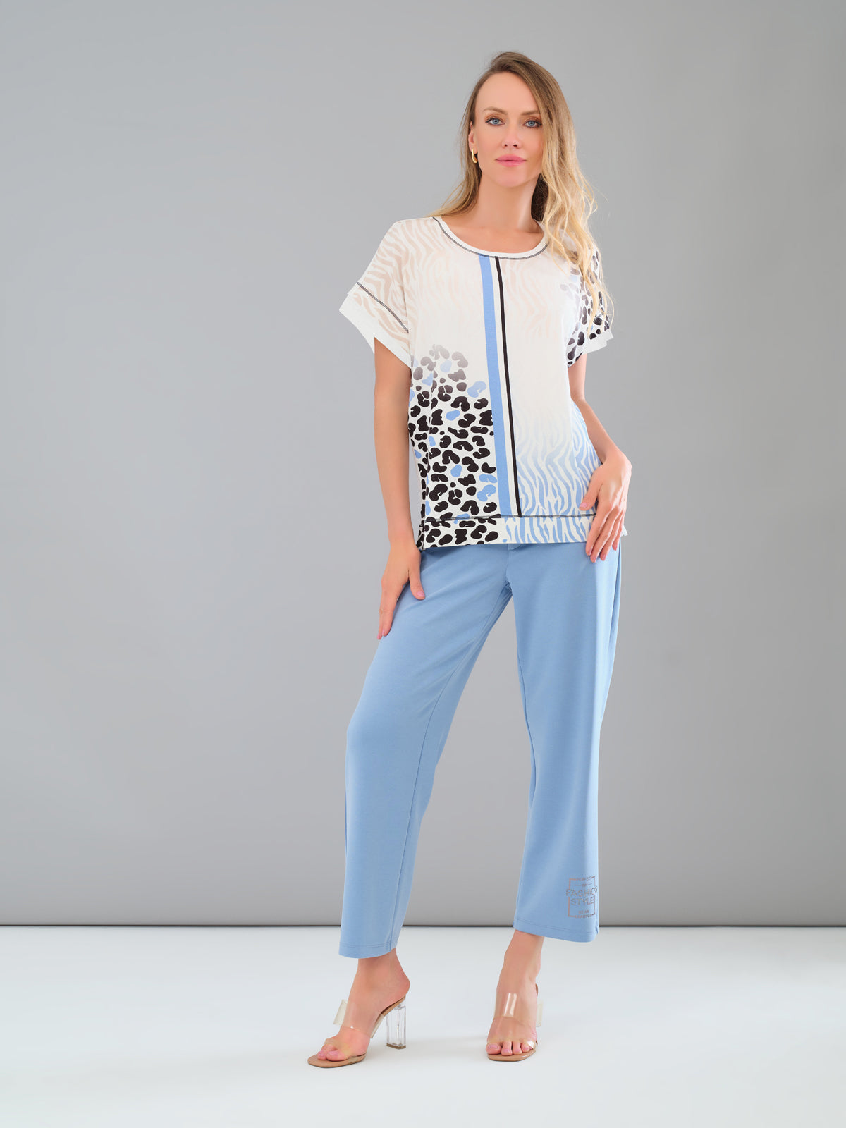 Woman wearing a stylish outfit with a patterned top and light blue pants on a gray background