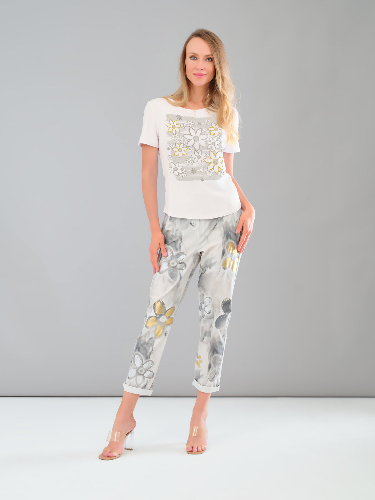 Woman wearing a white top with floral embellishments and floral pants on a gray background