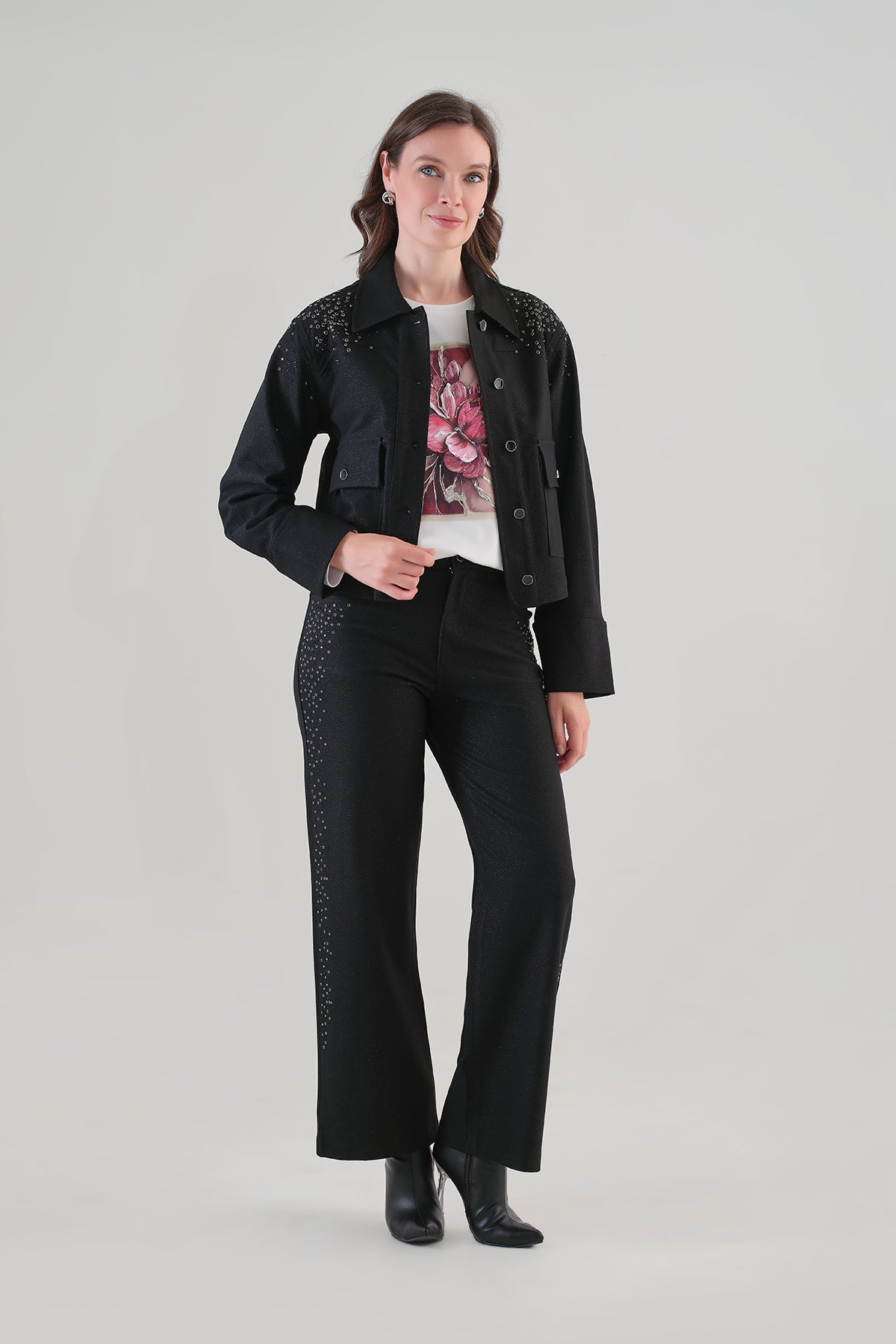A woman modeling a black cropped jacket with silver rhinestone embellishments on the shoulders and sides, paired with black wide-leg trousers, a white graphic t-shirt, and black ankle boots, against a light gray studio background.