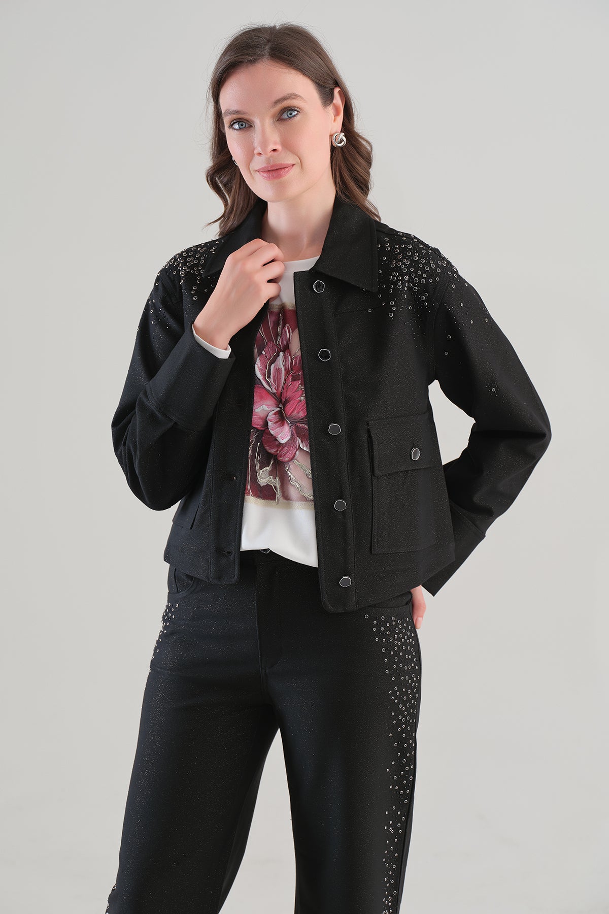 A woman modeling a black cropped denim jacket with silver rhinestone embellishments on the shoulders and sides, worn over a white graphic t-shirt and black pants, against a light gray studio background.