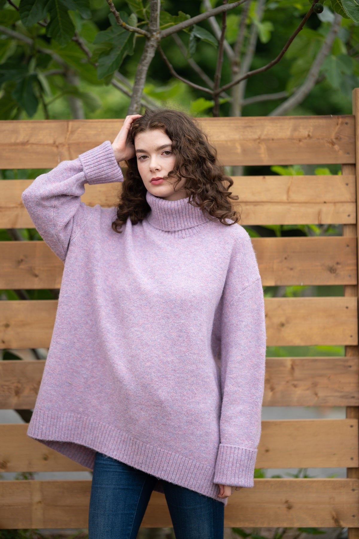 Woman wearing a light purple sweater in front of a wooden fence with greenery.