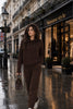 A woman walks down a wet city street wearing a matching dark brown lounge set consisting of a pullover hoodie and wide-leg sweatpants. She is carrying a brown handbag and wearing white sneakers.