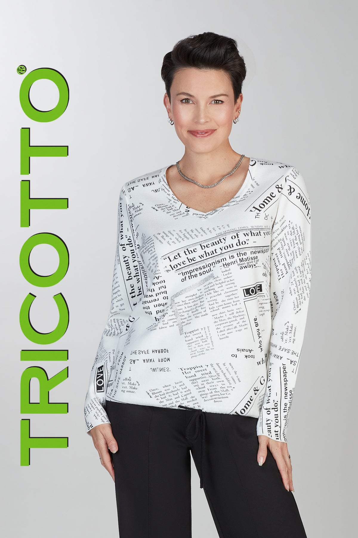 Woman wearing a white long-sleeve shirt with text pattern and black pants, standing against a gray background with 'Tricotto' branding.