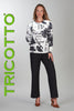 Woman wearing a black and white patterned top with black pants, standing against a gray background with 'TRICOTTO' branding.