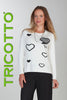 Woman wearing a white sweater with black heart patterns, standing against a plain background with 'TRICOTTO' branding.