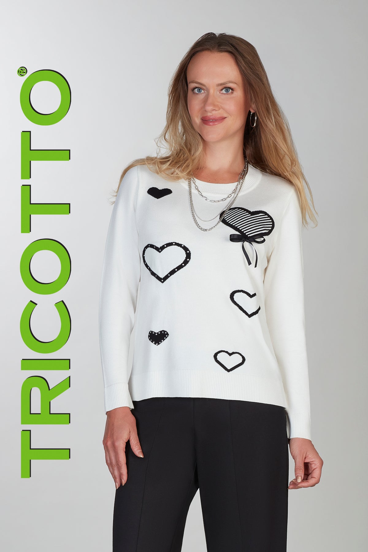 Woman wearing a white sweater with black heart patterns, standing against a plain background with 'TRICOTTO' branding.